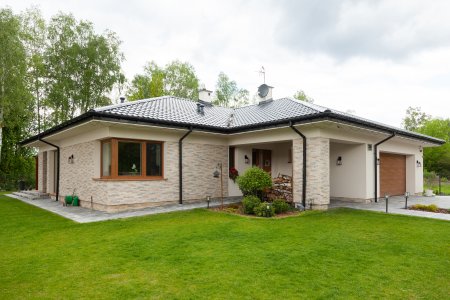 Gotowy dom parterowy z jasną elewacją z kamienia dekoracyjnego, ciemnym dachem i zintegrowanym garażem w otoczeniu zieleni.